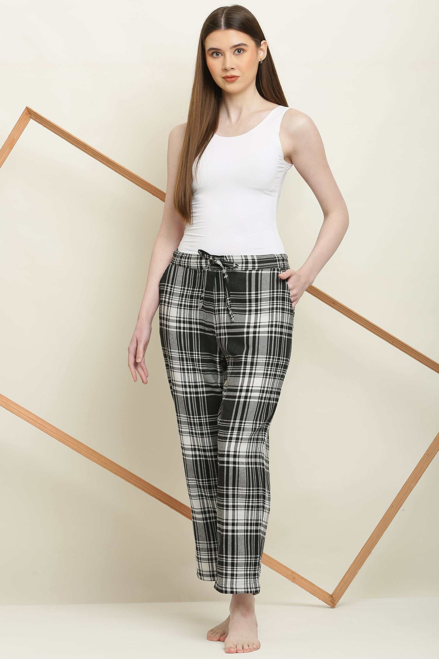 Woman wearing black and white checked cotton pyjama pants with elastic waistband and drawstring, paired with white tank top, standing barefoot.