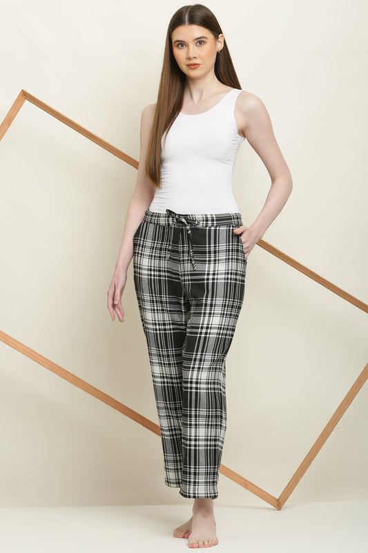 Woman wearing black and white checked cotton pyjama pants with elastic waistband and drawstring, paired with white tank top, standing barefoot.